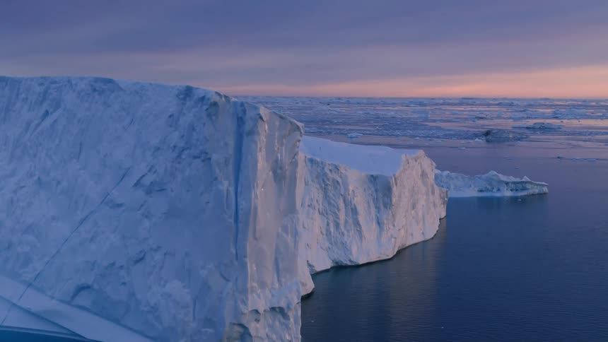 The video features an impressive aerial view of a massive towering iceberg with sheer vertical white and bluetinged ice walls dropping into the dark ocean water the scene is bathed in the soft twilight colors of the arctic sky with the horiz