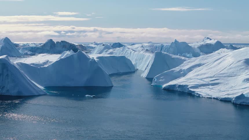Massive ice blocks float in a blue body of water the frozen structures show steep cliffs and snowy slopes the water opens into a channel between the formations and a bright sky with scattered clouds extends over the horizon