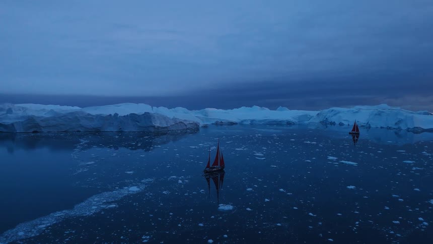 Two small boats with red sails moves across dark water surrounded by floating ice and massive white ice formations under a deep blue clouded sky creating a long reflection on the still water