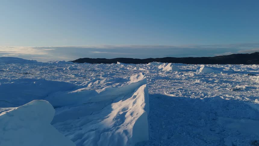 An aerial view slowly pulls back over a massive field of fractured snowcovered ice and broken icebergs extending towards the horizon the bright cold scene is set under a clear blue sky with a dark distant landmass visible in the background