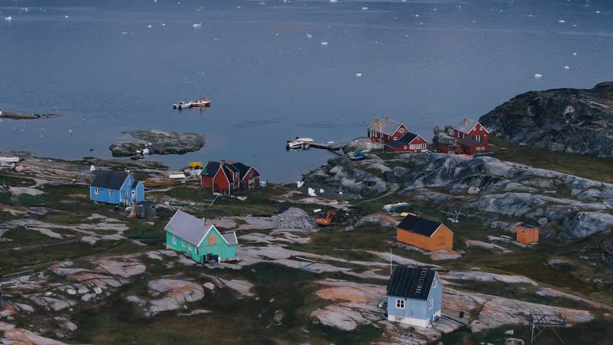 The video is a wide aerial shot of a small coastal town likely in greenland nestled among rocky terrain and dark waters scattered across the brown and green rocks are several small brightly colored houses including blue red and mint green str