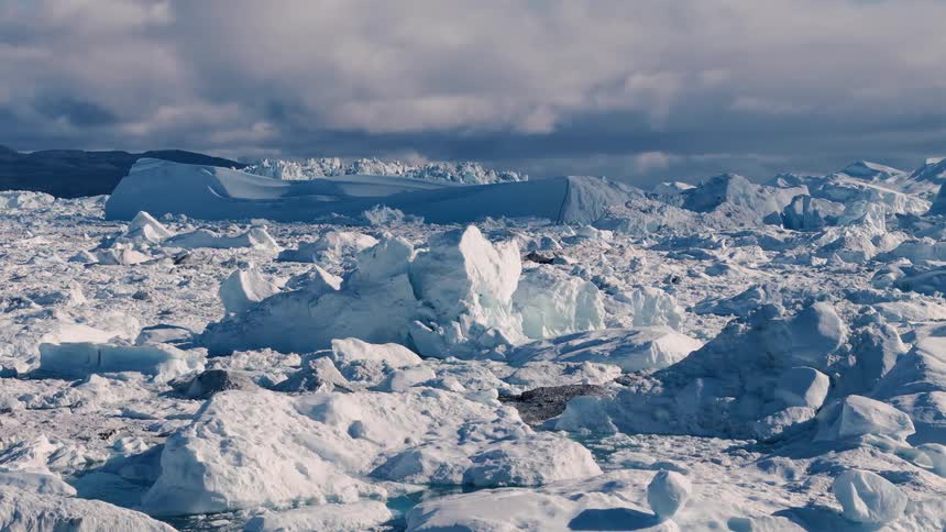 A vast collection of ice fragments and large ice formations floats in water under a sky with low clouds with a background of a landmass covered in snow and ice