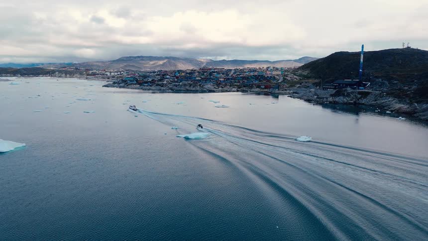 An aerial camera shot shows a small boat moving across a cold water body leaving a visible wake passing icebergs with a cluster of colorful buildings on the rocky shore in the distance framed by dark hills and a cloudy sky
