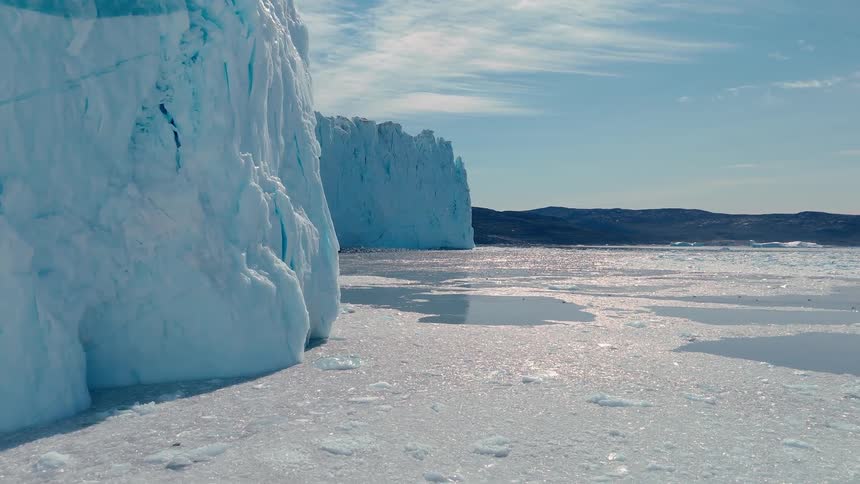 Bright towering walls of blue glacier ice meet a large expanse of floating ice and water under a sunny sky with distant mountains on the horizon