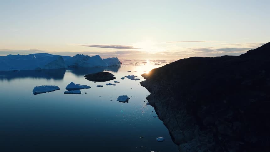 The video is an elevated shot of a glacial area in soft light where a dark rocky silhouette of a hill or mountain meets the water numerous small white icebergs float in the calm deep blue water with a bright sunset visible over the horizon