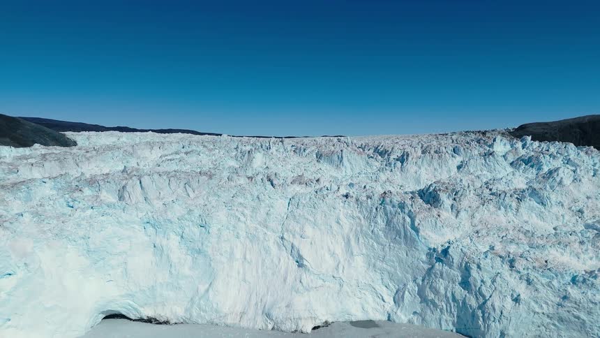 The video is a sweeping aerial shot of a massive heavily crevassed glacier face under a bright blue sky the textured white and blue ice descends from dark rocky slopes on either side of the frame