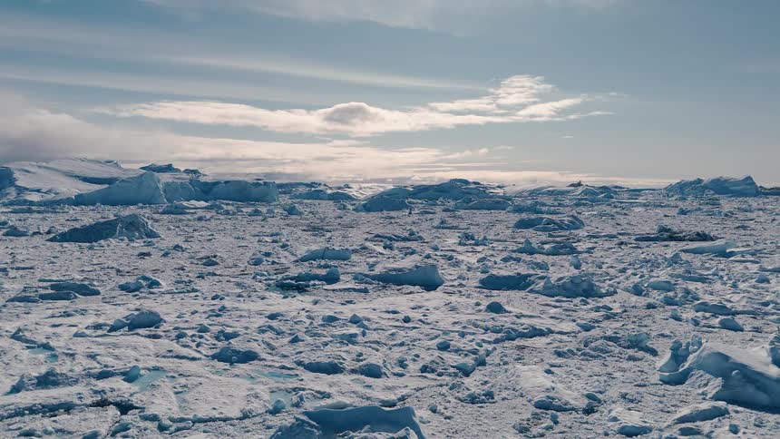 A vast flat area of broken white and blue ice extends toward the horizon under a pale sky with scattered white clouds bordered by large ice formations in the distance