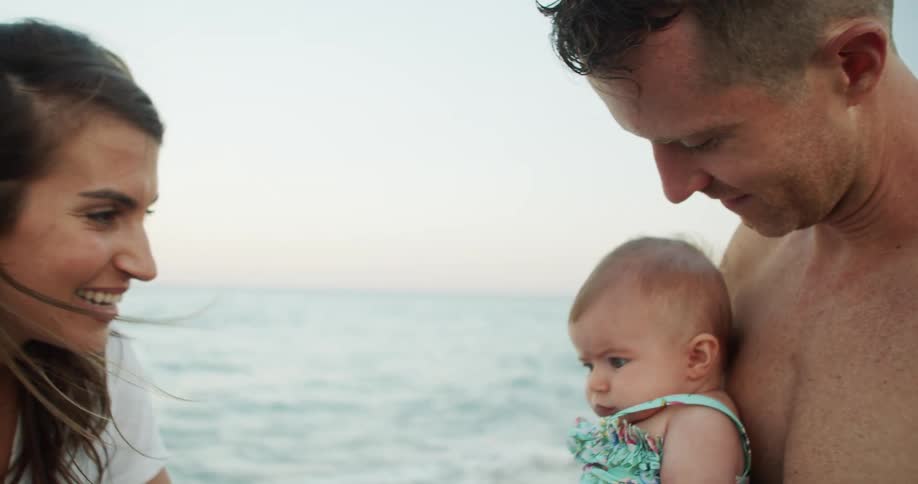 A close view of a shirtless man holding a baby in a swimsuit on a beach with a smiling woman in a white top looking at the baby in the background as waves crash onto the shore
