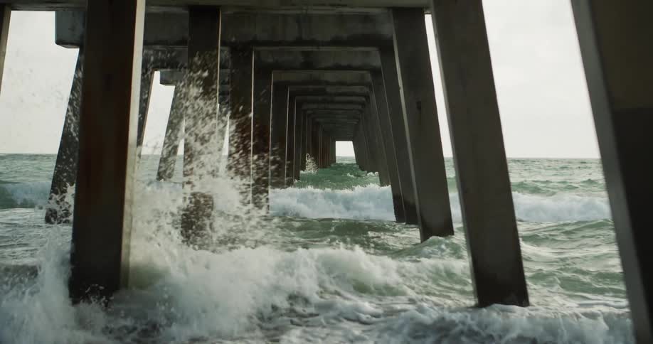 A view beneath a concrete pier shows waves breaking around the tall pillars as the sea surges in