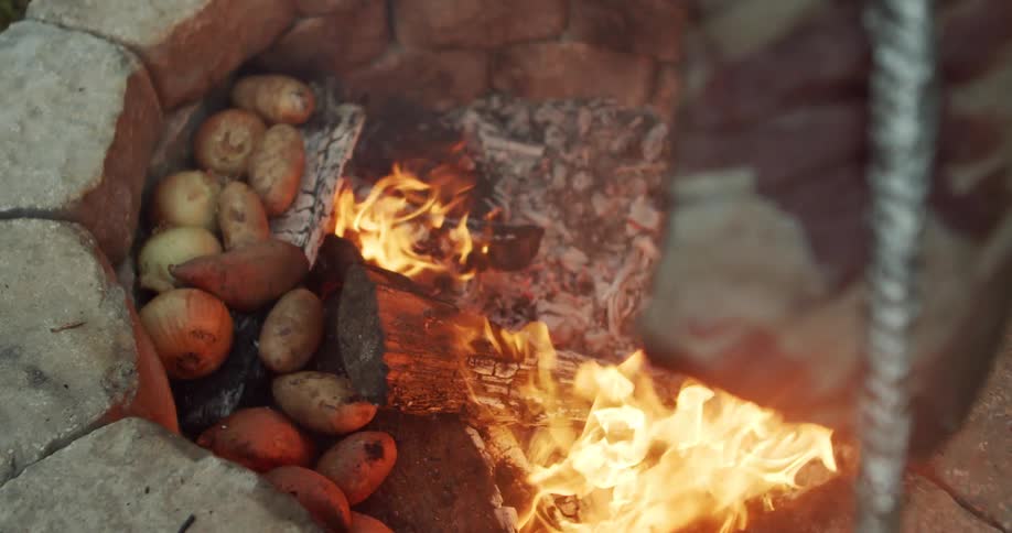 The video shows a closeup of potatoes onions and sweet potatoes being roasted over an open fire in a makeshift outdoor oven made of stonesthe vegetables are arranged around the edge of the pit while wood logs burn in the center cooking the