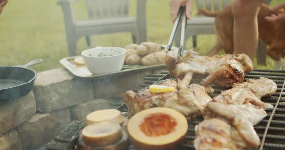 A person is outdoors using tongs to flip whole chickens and pieces of meat cooking over an open fire pit with a grill grate beside the fire a plate of potatoes and a sliced squash or pumpkin are visible while a dog wanders in the backgroun