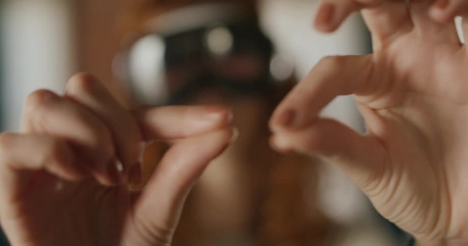 A person wears a virtual reality headset in the blurred background brings both hands into the foreground and uses thumbs and index fingers to perform a small pinching and spreading motion in the air