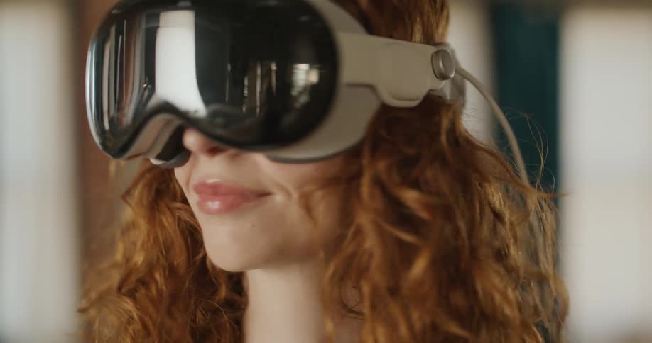 A woman with curly red hair wears a reflective vr headset smiles while looking forward moves her head slightly and briefly raises one hand toward the camera