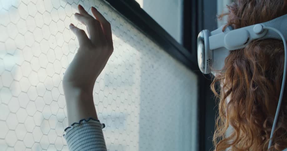 A blonde woman wearing a white virtual reality headset reaches out a hand to touch a window covered with a fine metal grid