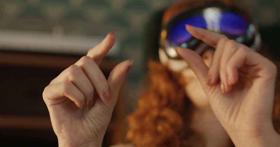 A closeup shot focuses on a young woman with curly red hair wearing a white virtual reality vr headset with bluetinted goggles her hands are held in front of her face performing a pinching or tapping gesture as she interacts with the virtual