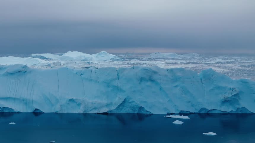 A broad ice wall floats in calm dark water beneath a heavy overcast sky with scattered floes on the horizon