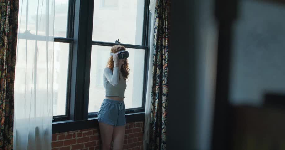 A camera shot shows a woman wearing a virtual reality headset standing indoors near a large window moving her hand in the air as if interacting with a display next to an exposed brick wall