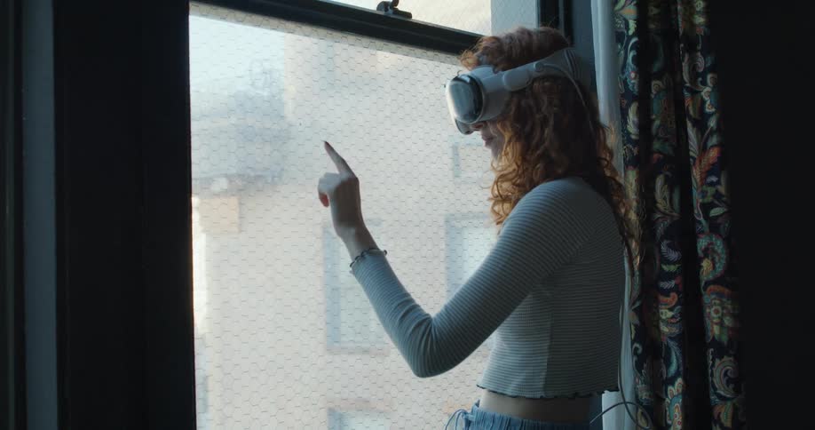 A young woman standing in front of a window wearing a virtual reality headset and moving her hand as if drawing something with her finger