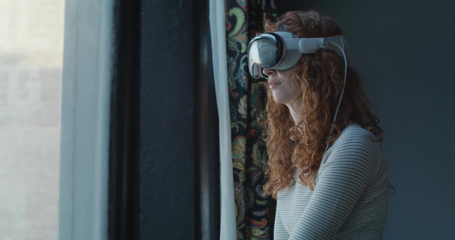 Teenage girl wearing a virtual reality headset stands near a window nodding her head