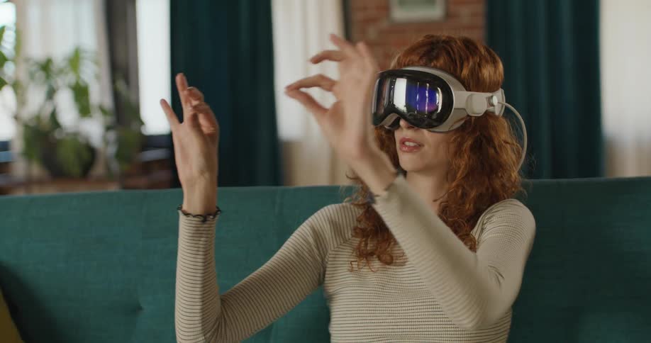 A woman with long curly red hair is sitting indoors and actively interacting with a virtual reality vr headset she is wearingshe is using her hands and fingers to make precise gestures and movements in the air suggesting she is manipulating