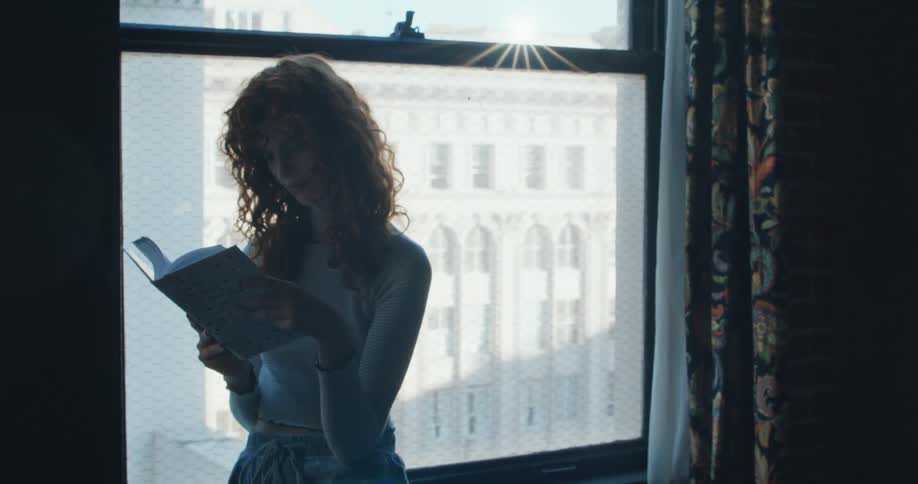 A young woman standing near a window reading a book