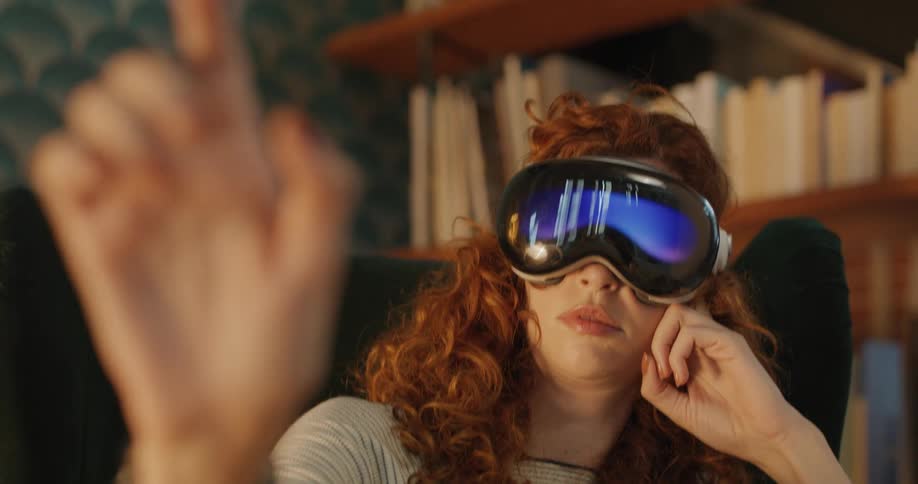 A woman with curly red hair sits in a chair wearing a vr headset and extends one hand into the foreground and rests her other hand on her face against a background of bookshelves in a indoor setting