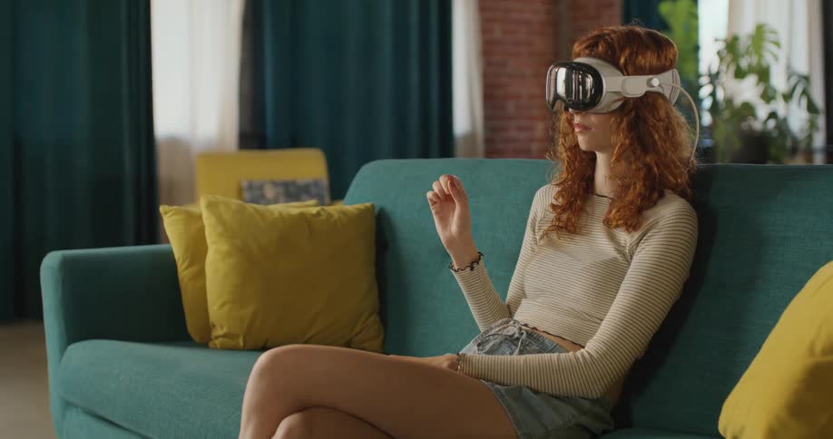 A young woman with curly red hair is seated on a teal couch wearing a white and gray virtual reality vr or mixed reality headset she is actively gesturing with her right hand in the air as if interacting with a digital interface that only she