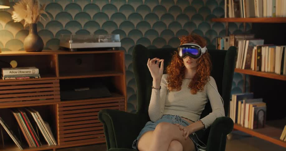 The video shows a woman with curly red hair wearing a white vrmr headset sitting in a dark green armchair in a room lined with wooden bookshelves and vintage wallpaper she is performing a hand gesture as if manipulating a virtual object or int