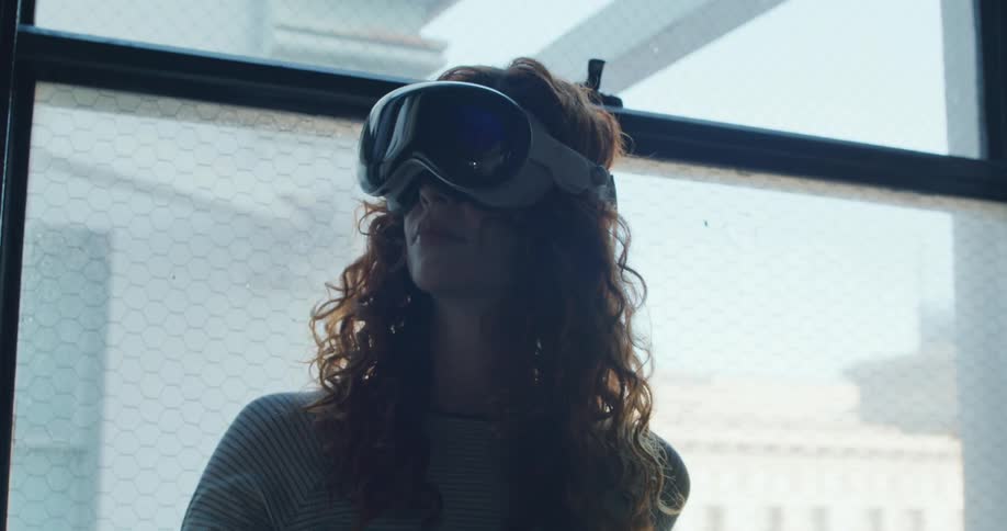A young woman standing near a window wearing a virtual reality headset and moving one of her hands to select something