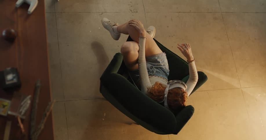 The video features a highangle shot of a young person with curly red hair sitting back dramatically in a dark green armchair indoors they are relaxing with their knees bent and hands raised in the air towards the ceiling