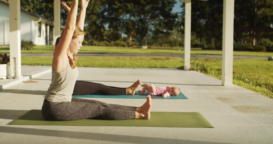 Two women sit on yoga mats with their legs extended and arms raised over their heads on a porch with a baby lying on a mat beside them a house and lawn are visible in the background under bright sunlight
