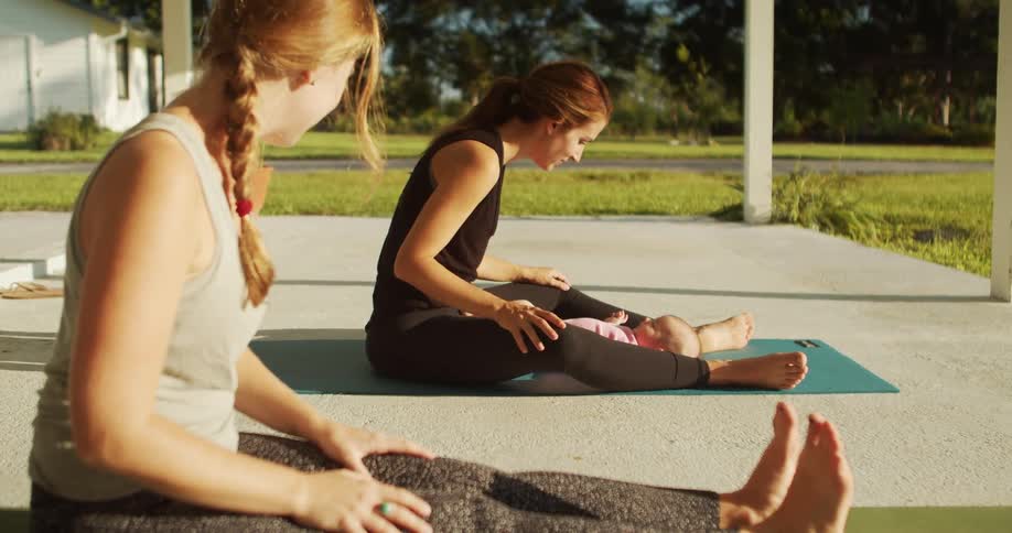Two young women sit outdoors on a yoga mat on the front porch with one holding an infant in her lap and playing with the baby