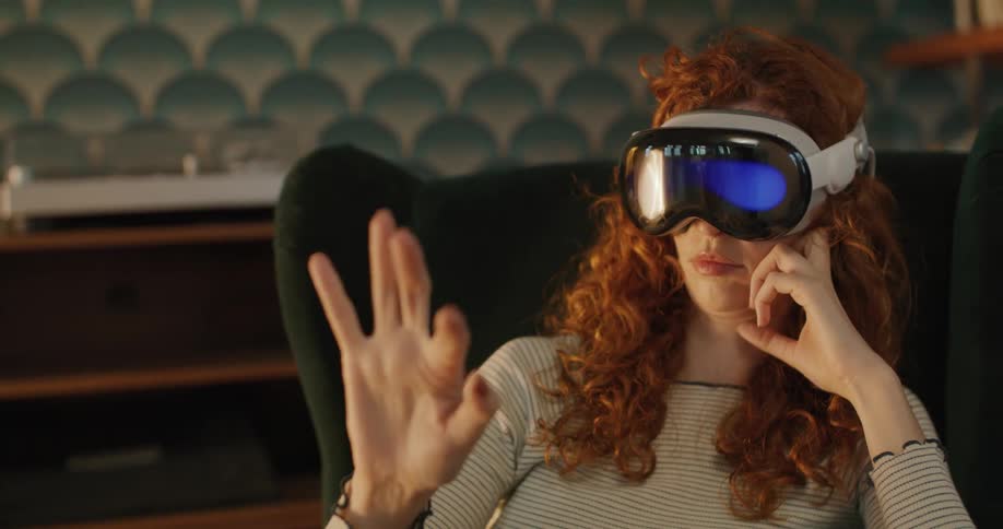 A woman with curly red hair sitting in an armchair wearing a white virtual reality headset with a dark visor glowing blue moving her hand in the air to interact with unseen virtual content