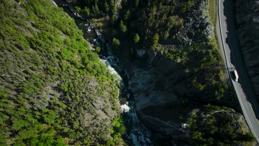 The video shows a high aerial view looking straight down into a deep narrow rocky gorge or canyona river with whitewater rapids flows through the base of the gorge and a road runs along the top edge of the righthand canyon wall with