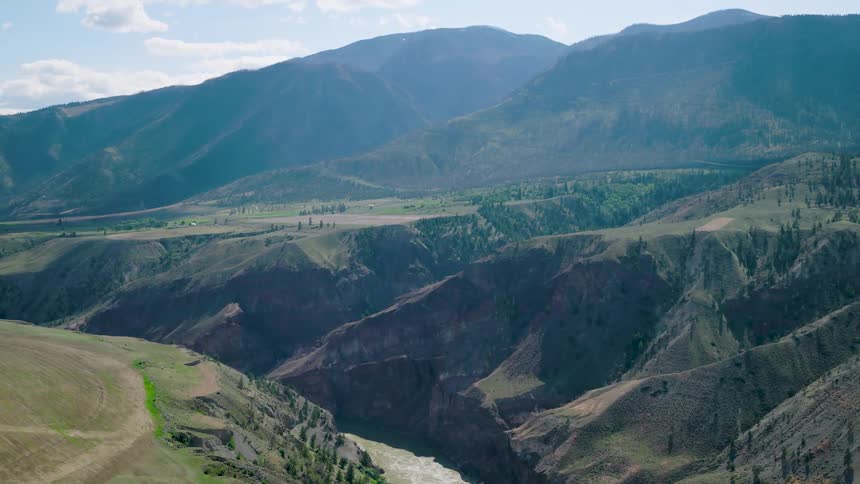 A muddy river flowing through a deep canyon with rolling green and brown hills leading to a vast mountain range under a partly cloudy sky