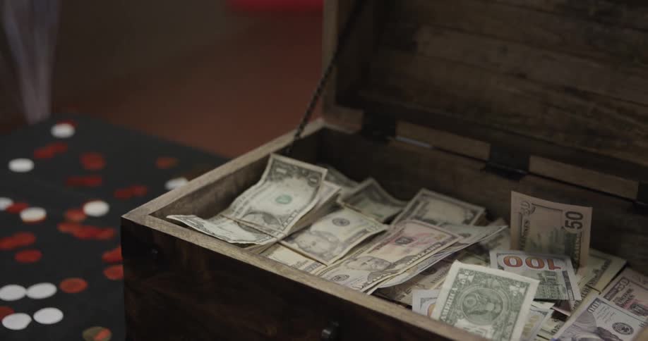 Two hands exchange paper money near a bundle of red balloons a person then moves the money toward a wooden box filled with various denominations of united states currency and places the bills inside the container