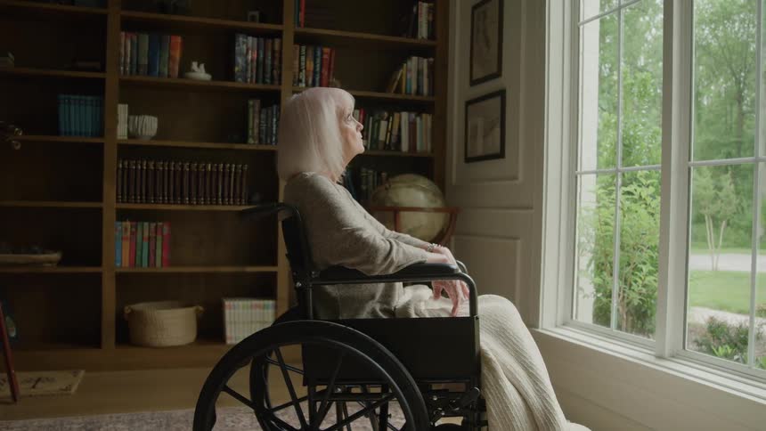 An elderly woman with white hair sits in a wheelchair wrapped in a blanket looking out a large window at a green lawn with a full wooden bookshelf behind her