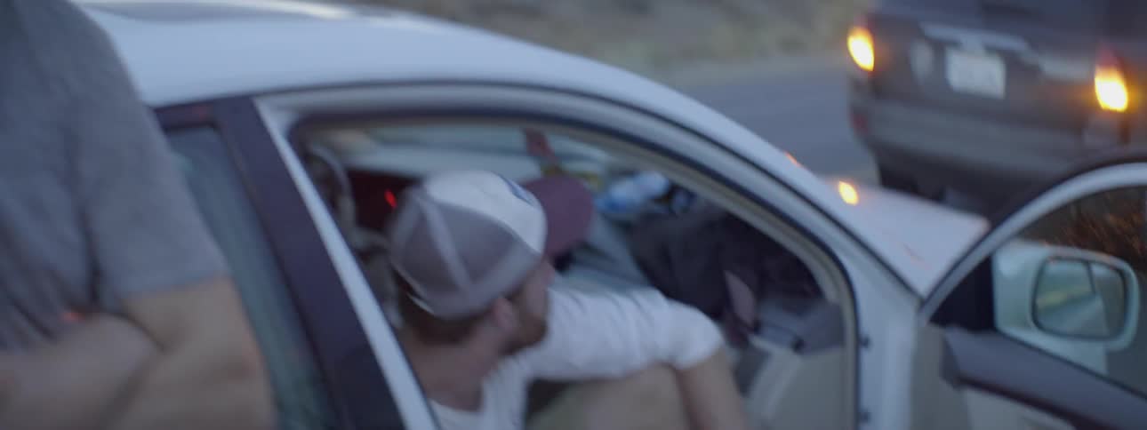 Two people sit and stand near a parked car