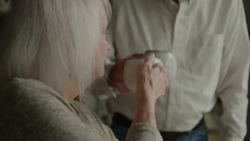 A close side view shows an elderly woman with white hair sitting in a wheelchair and sipping from a mug as an elderly man in a white shirt and jeans walks up behind her