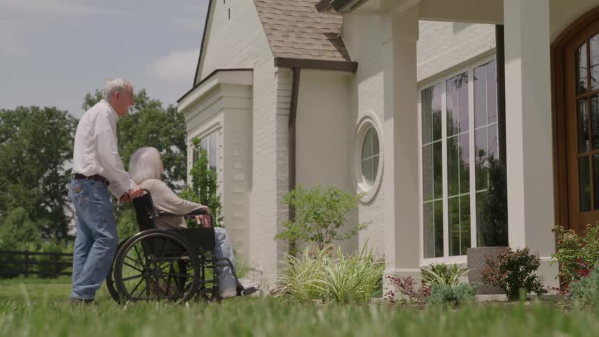 The video shows a lowangle shot of a man wearing a white shirt and jeans pushing an elderly woman in a wheelchair past the exterior of a white brick housethey are moving from the foreground toward the left passing a landscaped garden bed and