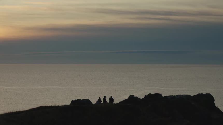 Three people sit on a promontory along the coast overlooking the water