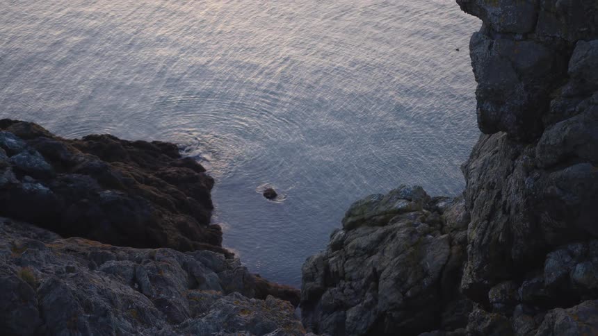 A narrow inlet of calm water is framed by rugged gray cliffs and rocky ledges from an overhead viewpoint