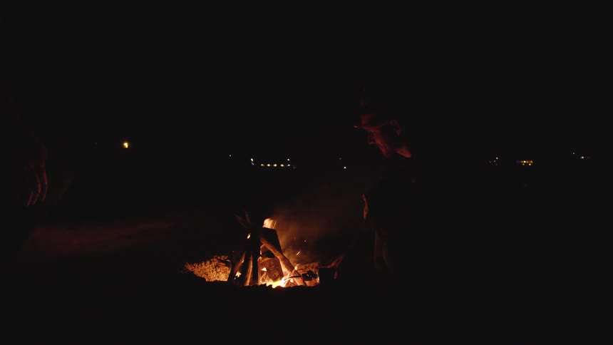 A fire burns in a pit on the ground at night illuminating the lower bodies of two silhouetted people standing nearby and one person adjusts the wood in the flames while smoke and sparks drift upward into the dark air