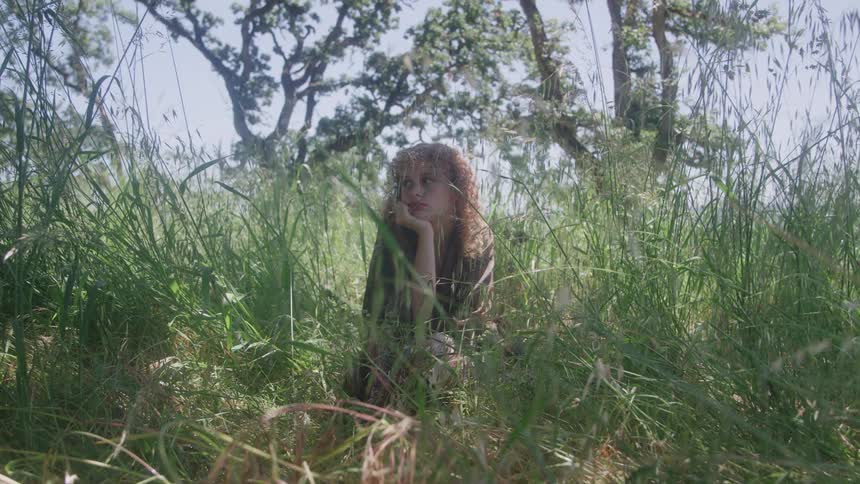 A teenager sits in tall green grass outdoors under bright sky