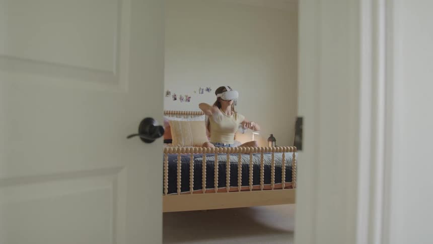 A wide view through a slightly open door shows a teenager sitting on her bed in a bright bedroom wearing a white virtual reality headset and playing with controllers