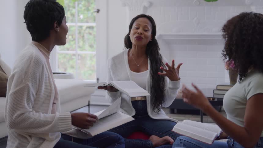 Three women are sitting on the floor in a bright white living room reading books during a casual gathering with the woman in the center speaking and gesturing with her hands