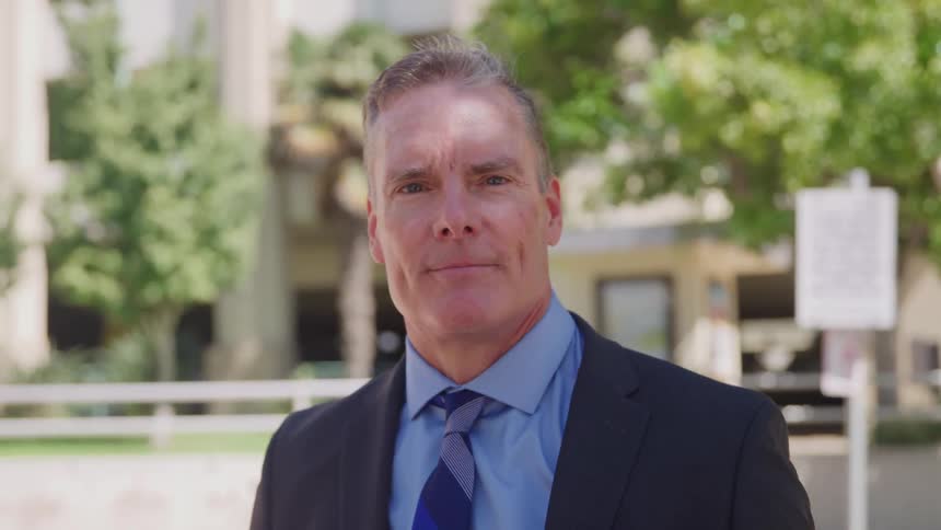 A close view shows a serious mature man wearing a black suit and a blue tie standing outdoors in front of a blurred background of green trees and buildings