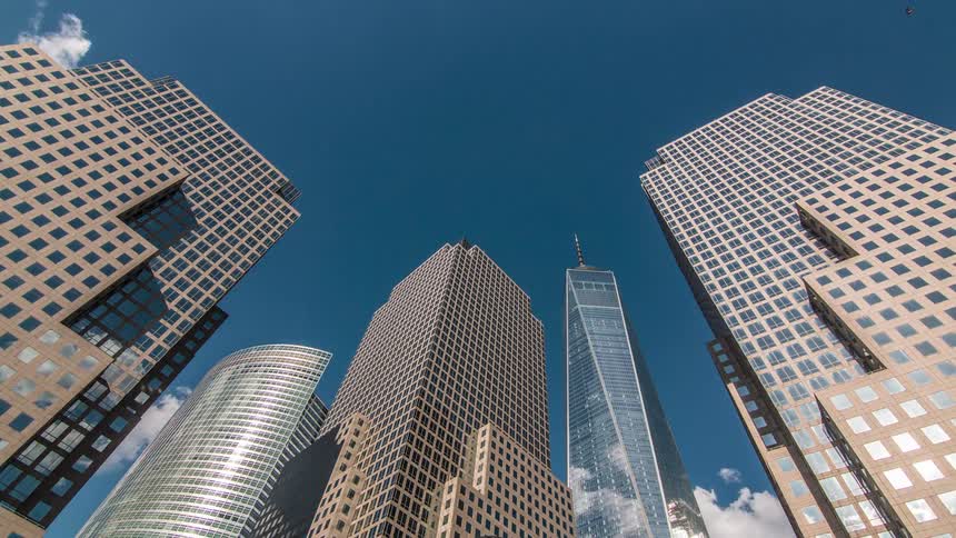 Several tall buildings with glass and stone exteriors rise vertically into a clear blue sky with white clouds filmed from a low position