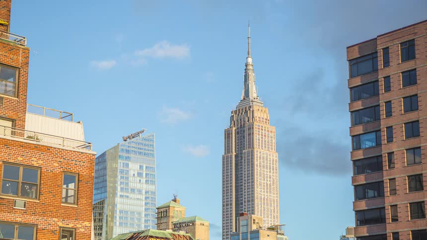The tall stepped central skyscraper rises toward a spire against a blue sky with white clouds flanked by a curved glass tower and two brick buildings on a clear day
