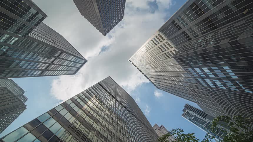 A dramatically angled low view from the ground looks up at the towering glass and steel facades of multiple modern skyscrapers against a bright blue and cloudy sky